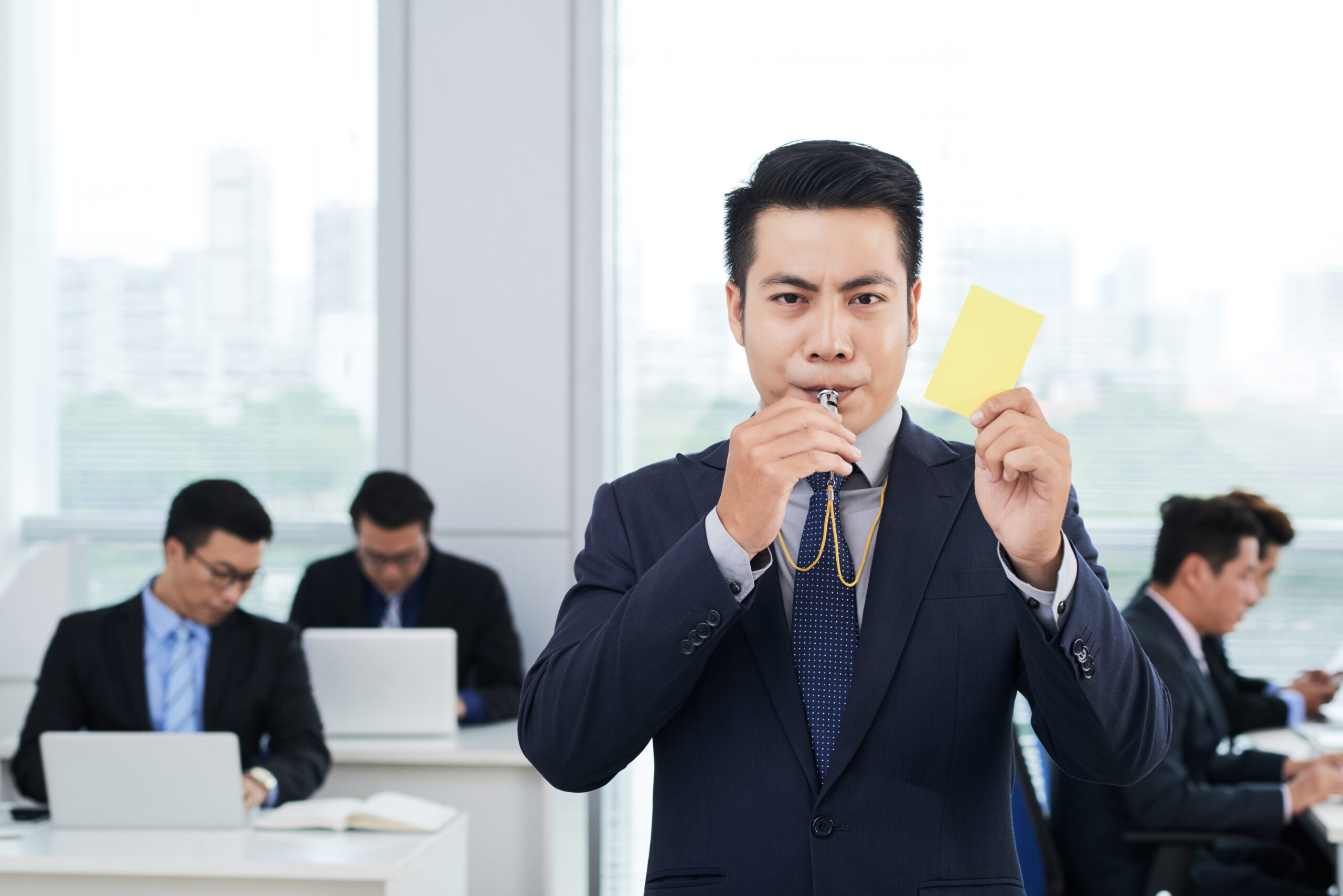 Waist-up portrait of handsome Asian entrepreneur wearing classical suit blowing whistle and showing yellow card to camera, interior of busy open plan office on background